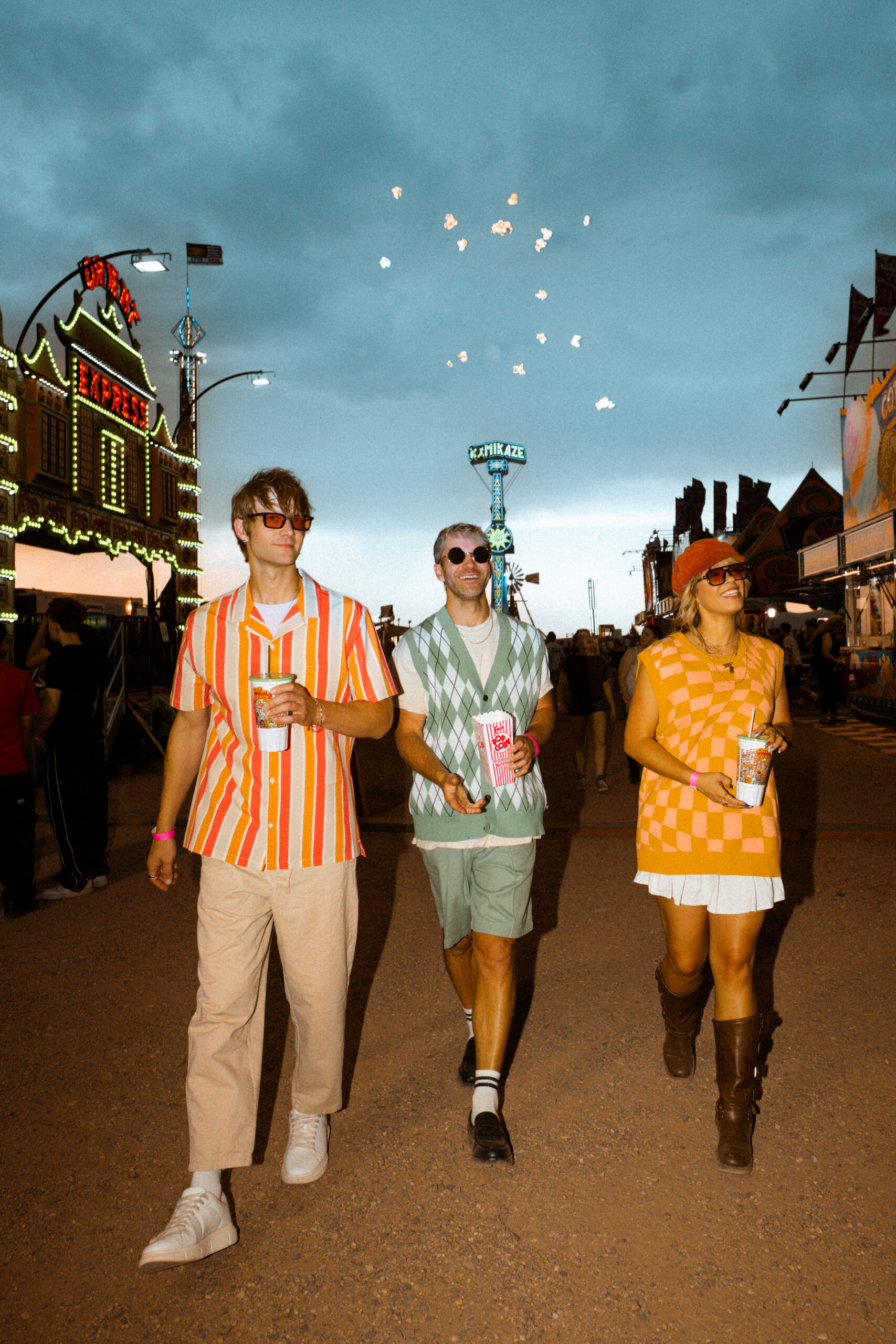 Three retro-styled friends walking through the fair midway with drinks and popcorn